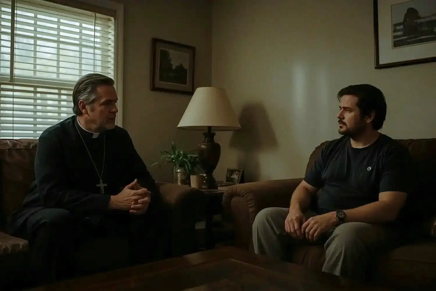 A priest in black clerical attire sits on a couch, engaging in a conversation with a man in a dark shirt, in a warmly lit living room with a lamp and framed pictures.